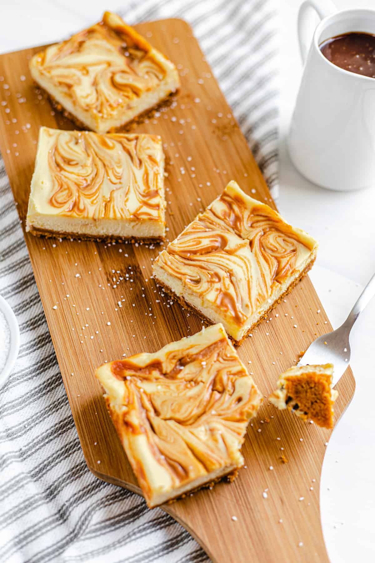 cheesecake bars on a wooden board with pitcher of caramel next to it and bite taken out of the front bar