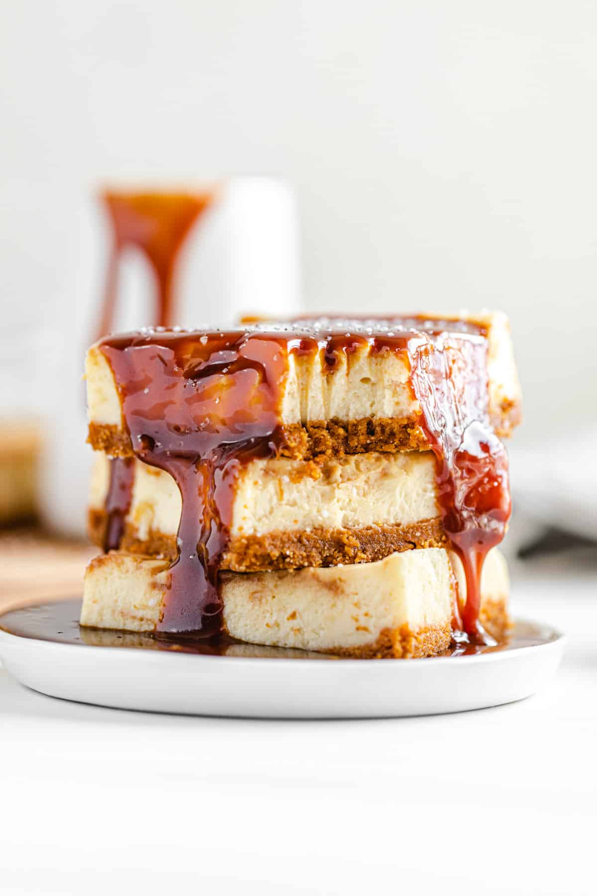 stack of cheesecake bars on a small white plate with a pool of caramel sauce on it and a pitcher of caramel in the background