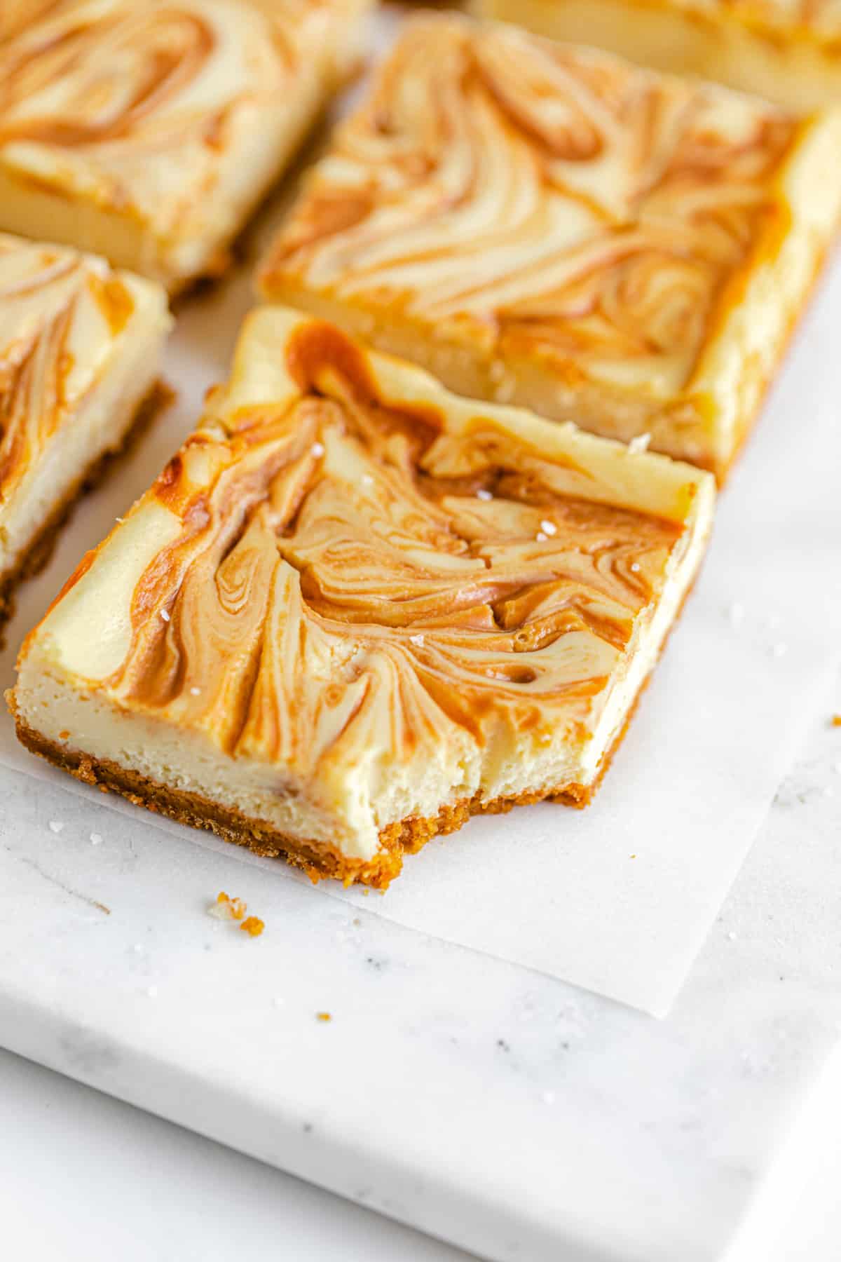 close up of cheesecake bars with caramel swirled on top in rows on a square marble board and a bite taken out the front bar