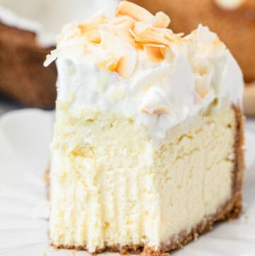 A close-up slice of cheesecake topped with whipped cream and toasted coconut flakes on a white plate, with a fork resting beside it and the whole cheesecake visible in the background.