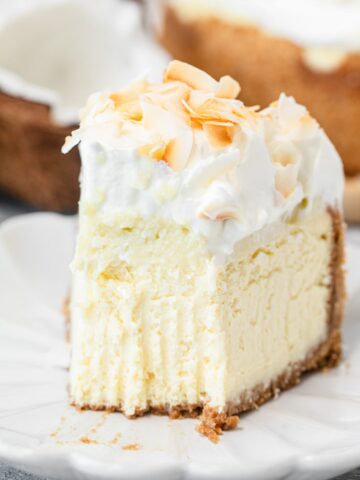 A close-up slice of cheesecake topped with whipped cream and toasted coconut flakes on a white plate, with a fork resting beside it and the whole cheesecake visible in the background.