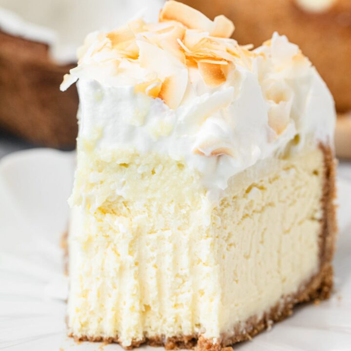 A close-up slice of cheesecake topped with whipped cream and toasted coconut flakes on a white plate, with a fork resting beside it and the whole cheesecake visible in the background.