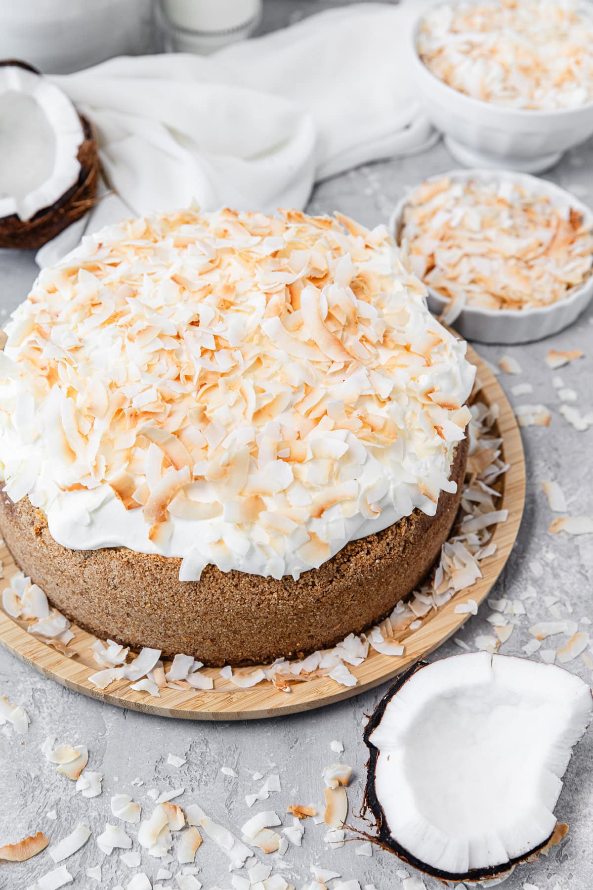 A whole cheesecake topped with toasted coconut flakes and whipped cream, displayed on a wooden plate with extra coconut pieces scattered around on a light gray surface.