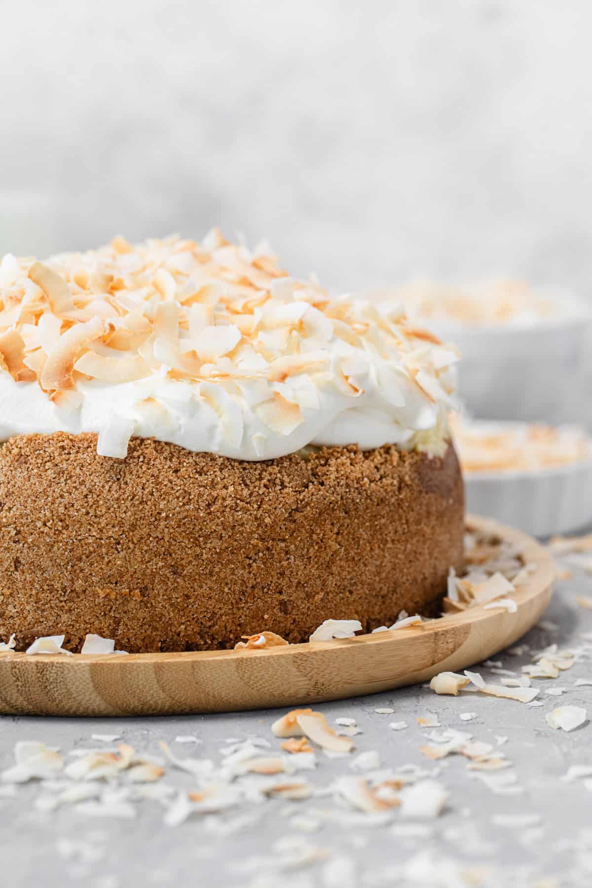 Close-up of a whole cheesecake topped with whipped cream and toasted coconut flakes on a wooden plate, with coconut pieces scattered around on a light gray surface.