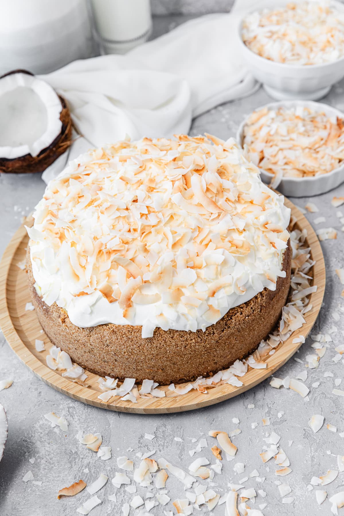 A whole cheesecake topped with toasted coconut flakes and whipped cream, displayed on a wooden plate with extra coconut pieces scattered around on a light gray surface.