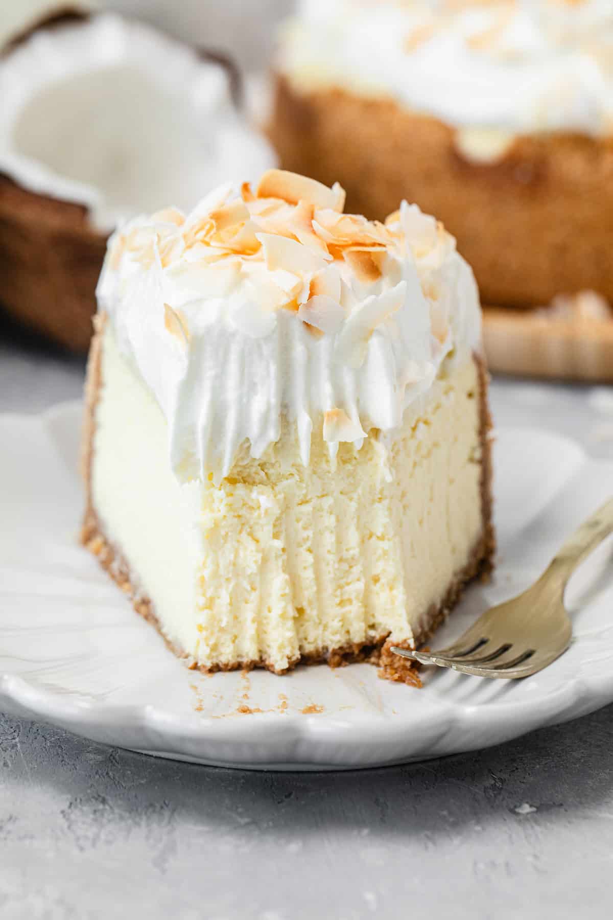 A creamy slice of cheesecake topped with whipped cream and toasted coconut flakes on a white plate, with a fork resting beside it and the whole cheesecake visible in the background.
